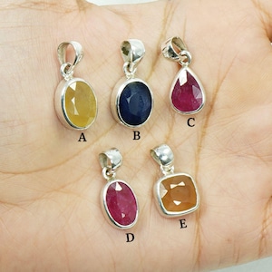 May include: Five gemstone pendants with silver settings. The gemstones are in various shapes: oval, teardrop, and square. The colours include yellow, blue, and red. The pendants are displayed on a hand.