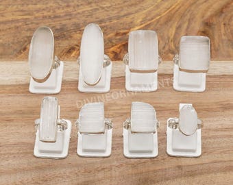 May include: Eight silver rings with white stone settings in different rectangular and oval shapes. The rings are displayed on white stands, set against a light brown wooden surface. The rings have a modern, minimalist design.