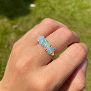 May include: A silver ring with five oval-shaped opals. The opals are iridescent with shades of blue, green, and pink.