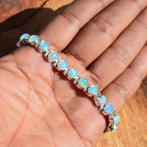 May include: A silver bracelet with a row of oval-shaped opal gemstones. The bracelet is on a hand, and the opals have a light blue and green iridescent sheen.