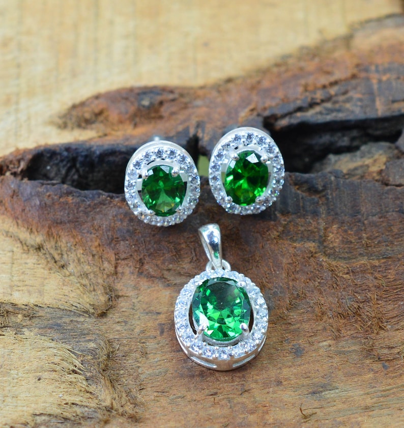 May include: A silver pendant and earrings set with a green gemstone surrounded by a halo of smaller white gemstones.