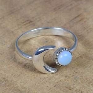 May include: A silver ring featuring a crescent moon design and a light blue opal gemstone. The ring is adjustable and can be worn on any finger.