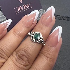 Könnte beinhalten: Ein silberner Ring mit einem grünen Edelstein, der in einem filigranen Design eingefasst ist. Der Ring wird an einem Finger mit weißer French Manicure getragen.