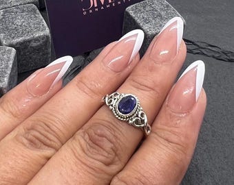 Puede incluir: Un anillo de plata con una piedra preciosa púrpura engastada en un diseño delicado y ornamentado. El anillo se lleva en un dedo con una manicura francesa blanca.