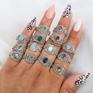 May include: A set of 12 silver rings with different colored gemstones. Each ring features a delicate, ornate design with a single oval-shaped gemstone.