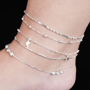 Puede incluir: Una colección de tobilleras plateadas, cada una con un diseño único. Las tobilleras presentan varios estilos, incluyendo diseños de cuentas, cadenas y dijes. Una tobillera tiene un pequeño dije con forma de delfín. Sobre un fondo negro.