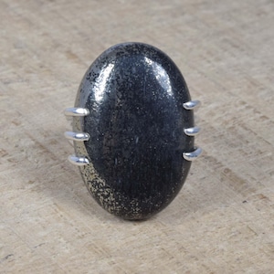 Puede incluir: Un anillo de plata con una gran piedra preciosa ovalada negra engastada en el centro. La piedra preciosa tiene un aspecto moteado y metálico.