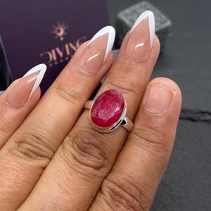 Könnte beinhalten: Ein silberner Ring mit einem großen, ovalen, roten Edelstein. Der Ring wird an einem Finger getragen.