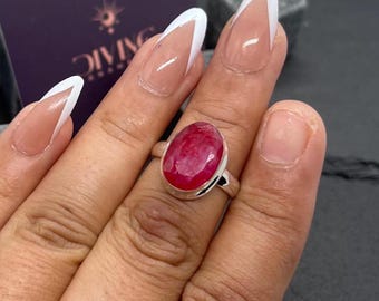 Puede incluir: Un anillo de plata con una piedra preciosa roja grande de forma ovalada. El anillo está en el dedo de una persona.
