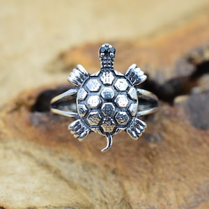 Puede incluir: Un anillo de plata con forma de tortuga con una piedra preciosa negra en la cabeza. El caparazón de la tortuga está detallado con un patrón texturizado.