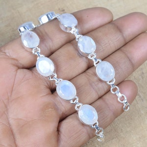 May include: A silver bracelet featuring oval-shaped, faceted moonstone gemstones. The gemstones are linked together with small silver connectors. The bracelet is displayed on a light brown surface.