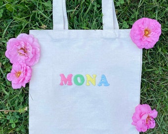Könnte beinhalten: Weiße Stofftasche mit dem Namen "MONA" in bunten Buchstaben bestickt. Die Tasche ist von rosa Blumen umgeben.