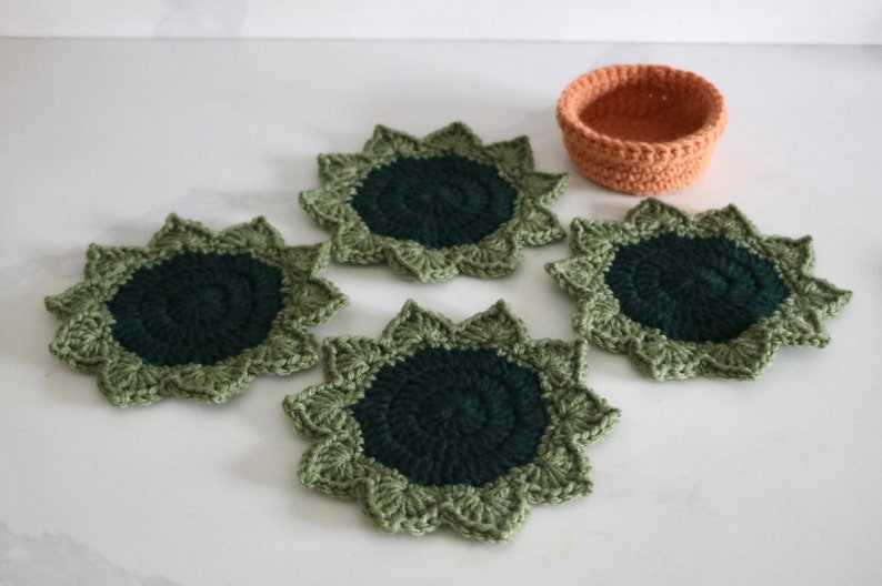 May include: Four green and dark green crocheted coasters with a starburst design. A smaller orange crocheted bowl is in the center of the image.