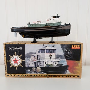May include: A black and green toy tugboat bank with the text "Texaco Fire Chief" on the side. The bank is on a stand and is in a box with the text "Texaco "Fire Chief" Tugboat Bank - First in a Series" on the front.