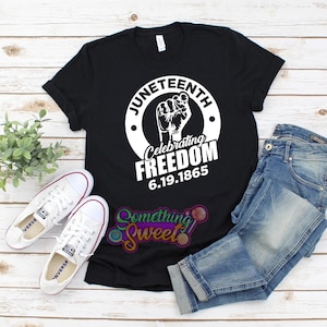 May include: Black t-shirt with a white graphic of a raised fist and the text "Juneteenth Celebrating Freedom 6.19.1865".