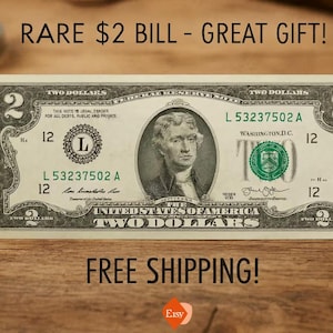 Pode incluir: Uma nota de dois dólares com um retrato de Thomas Jefferson, com o texto "RARE $2 BILL - GREAT GIFT!" no topo. A nota está sobre uma superfície de madeira. O texto "TWO DOLLARS" é visível.