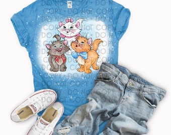 Puede incluir: Una camiseta azul con un diseño de lejía de tres gatos de dibujos animados. Los gatos son blanco, gris y naranja. El gato blanco tiene un lazo rosa, el gato gris tiene un lazo rojo y el gato naranja tiene un lazo azul.