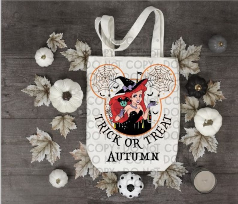 May include: White canvas tote bag with a Mickey Mouse silhouette design. The silhouette features a red-haired woman in a witch's hat and a black dress holding a potion bottle. The text "Trick or Treat Autumn" is printed below the silhouette.