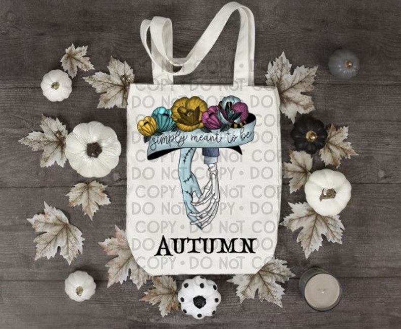 May include: A white canvas tote bag featuring a graphic of two skeletal hands holding each other, adorned with yellow, blue, and purple flowers. A banner above the hands reads "simply meant to be" and the word "AUTUMN" is printed below in black.