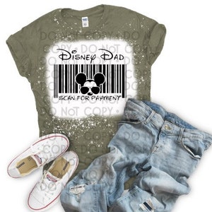 Könnte beinhalten: Ein olivgrünes T-Shirt mit einem weißen Used-Design mit dem Text "Disney Dad" über einem Barcode mit einem Mickey Mouse-Kopf mit Sonnenbrille. Das Bild enthält auch hellblaue Jeans und weiße Turnschuhe.