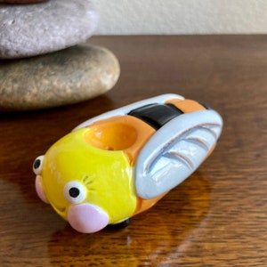 May include: A ceramic bee-shaped pipe with yellow, orange, black, and white paint. The pipe has a round hole in the top for smoking.