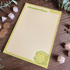 Könnte beinhalten: Ein rechteckiger Notizblock mit hellgelbem Papier, horizontalen Linien und dem Text Adventure Notes oben. In der unteren rechten Ecke befindet sich eine Illustration eines grünen gallertartigen Würfels mit Totenkopf, Schwert und Schild.