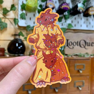 Op de afbeelding: Een sticker met drie cartoon-draken op elkaar gestapeld. De bovenste draak draagt een geel shirt en heeft een rood-oranje lichaam. De middelste draak is rood-orange en de onderste draak is rood-orange. De sticker heeft een glinsterende afwerking.