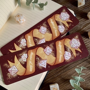 Könnte beinhalten: Zwei braune Lesezeichenkarten mit goldenen Banddesigns und weißen Würfeln mit goldenen Zahlen. Der Text auf dem Band lautet "Bildhauer von Geschichten" und "Meister des Schicksals".