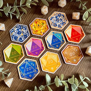 Può includere: Un set di dieci adesivi da dado a 20 facce con diversi design. Gli adesivi sono stampati su carta bianca e hanno un bordo dorato. I dadi sono numerati da 1 a 20. Gli adesivi sono disposti in cerchio su una superficie di legno.