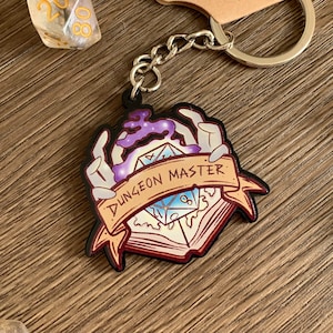 May include: A metal keychain featuring a purple and blue dice with the text "Dungeon Master" on a banner. The dice is surrounded by two hands reaching out from a book.