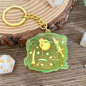 May include: A green slime keychain with a gold chain. The slime is filled with gold bones, a sword, and a skull. The keychain is on a wooden surface with white dice.