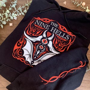 Può includere: Felpa con cappuccio nera con una stampa grafica rossa e bianca di un pipistrello con gli occhi e il testo "The Nine Hells".