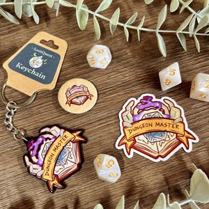 Könnte beinhalten: Eine Sammlung von Gaming-Zubehör, darunter ein Schlüsselanhänger, Aufkleber und Würfel. Der Schlüsselanhänger hat ein Etikett mit der Aufschrift "-LootQuest- Keychain". Die Aufkleber und der Schlüsselanhänger zeigen ein "Dungeon Master"-Design. Die Würfel sind weiß mit goldenen Zahlen.