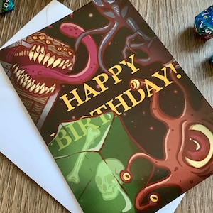 May include: A birthday card with a dark background and a colorful illustration of a monster with multiple eyes and a long tongue. The card says "Happy Birthday!" in yellow letters.