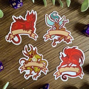May include: Four die-cut stickers featuring fantasy creatures, including a red dragon, a blue-winged fairy, a green horned imp, and a red winged demon. Each creature is positioned above a blank, light-tan ribbon banner for custom text.