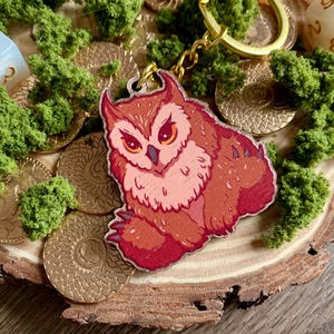 Può includere: Un portachiavi in legno con un'illustrazione stilizzata di un gufo marrone e rosso con grandi occhi arancioni, orecchie a punta e artigli affilati. Il portachiavi è fissato a un anello e a una catenella in metallo color oro.
