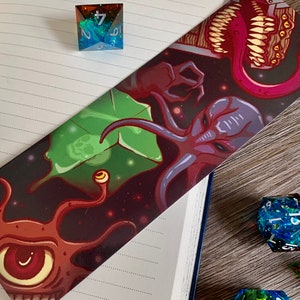 May include: A bookmark with a dark background and a colorful illustration of three space creatures. The creatures are a purple and red octopus-like creature, a green plant-like creature, and a brown creature with a large eye and multiple tentacles. The bookmark is surrounded by dice.