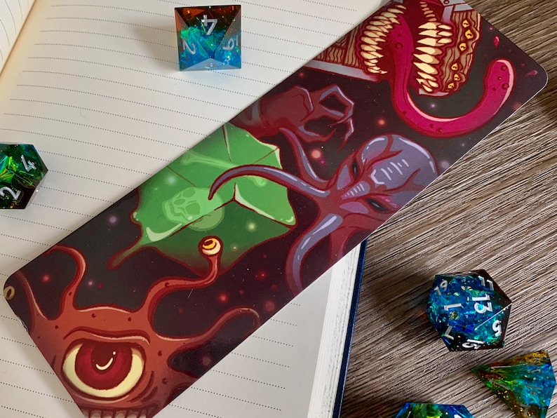 May include: A bookmark with a dark background and a colorful illustration of three space creatures. The creatures are a purple and red octopus-like creature, a green plant-like creature, and a brown creature with a large eye and multiple tentacles. The bookmark is surrounded by dice.