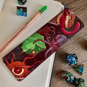 May include: A bookmark with a dark background featuring a colorful illustration of a tentacled creature with a large eye, a green leaf, and a red tongue with teeth. The bookmark is lying on a notebook with a pen and dice.