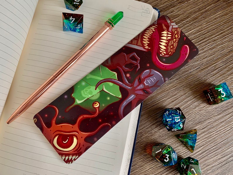 May include: A bookmark with a dark background featuring a colorful illustration of a tentacled creature with a large eye, a green leaf, and a red tongue with teeth. The bookmark is lying on a notebook with a pen and dice.