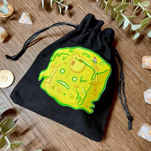 May include: Black drawstring dice bag with a yellow and green cartoon design. The design features a skull, sword, and other elements. Several dice and tokens are scattered around the bag on a wooden surface.