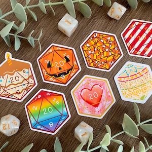 May include: A collection of seven hexagonal stickers featuring twenty-sided die designs with various patterns including a birthday cake, a jack-o'-lantern, candy corn, red stripes, a rainbow gradient, a red heart, and a decorative floral motif.