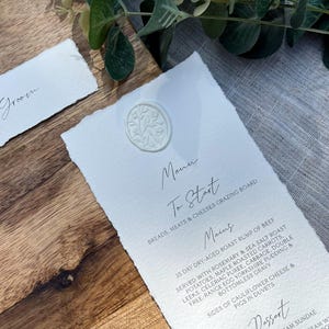 Puede incluir: Una tarjeta de menú de boda blanca con un sello de cera sobre una superficie de madera. El menú incluye una tabla de quesos y embutidos, carne asada y un sundae de helado.