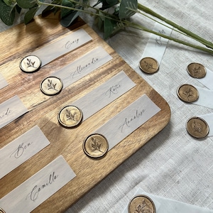 Puede incluir: Una tabla de madera con tarjetas de lugar de papel blanco y sellos de cera dorada. Las tarjetas de lugar tienen nombres escritos en caligrafía, incluyendo "Groom", "Bride", "Ben", "Camille", "Alexendre", "Katie", "Annalise", "Greg" y "Pen".