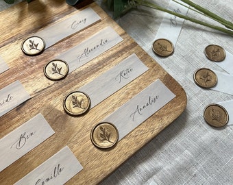 Può includere: Un tagliere di legno con cartoncini per segnaposto bianchi e sigilli di cera dorata. I segnaposto hanno nomi scritti in calligrafia, tra cui "Groom", "Bride", "Ben", "Camille", "Alexendre", "Katie", "Annalise", "Greg" e "Pen".