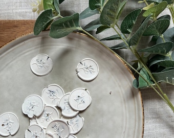 Puede incluir: Un plato blanco con un montón de sellos de cera blanca con un diseño floral. Los sellos están esparcidos por el plato. El plato está sobre un mantel de lino blanco. Una rama de eucalipto está en el fondo.