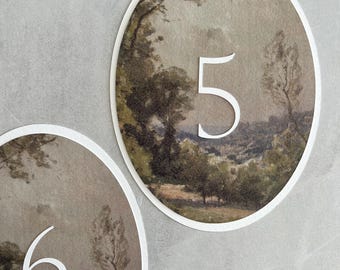 May include: Two oval table number cards. Each card features a landscape painting with a white number. One card displays the number 5, and the other displays the number 6. The background is a light gray.
