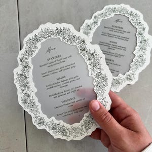 May include: Two white, floral-edged menu cards with a gray center. The cards feature a menu with starters, mains, and desserts. The menu includes items like roasted tomato soup and goat cheese salad.