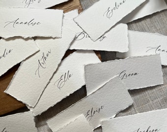 Könnte beinhalten: Eine Sammlung weißer, rechteckiger Platzkarte mit handschriftlichen Namen in schwarzer Tinte. Die Namen umfassen George, Annalise, Arthur, Ella, Eloise, Craig, Evelyn, Groom, Jackson und mehr.