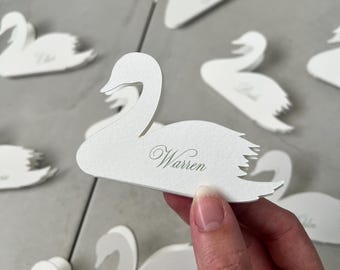 May include: White swan-shaped place cards with names printed in a delicate script. The cards are arranged on a light gray surface. One card is held up, displaying the name "Warren". These elegant cards are ideal for weddings or special events.